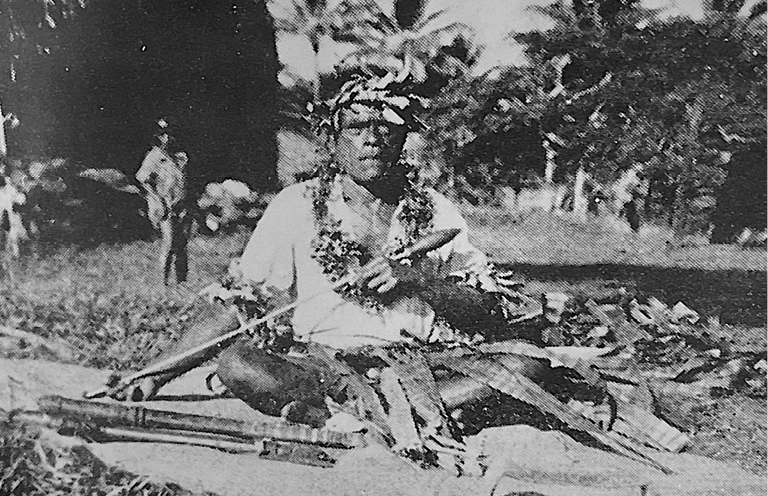 A black and white photograph of a Polynesian storyteller, seated on the ground with traditional adornments. He holds a wooden object, possibly a musical instrument or ceremonial item, while other people stand in the background.