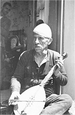 A historical black and white photograph of an elderly man sitting outside, playing a traditional stringed musical instrument. He appears to be engaged in an expressive performance, likely a folk musician.