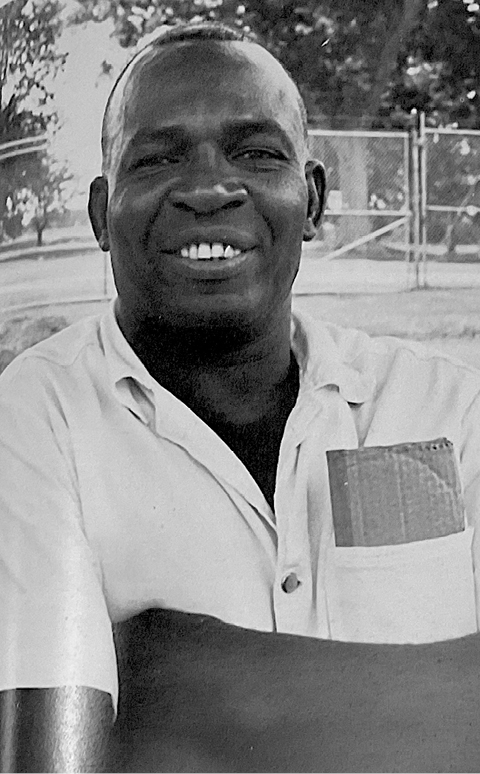A black and white photograph of a smiling man, wearing a short-sleeved shirt. His relaxed posture and warm expression suggest a candid and friendly moment, possibly a musician or storyteller.