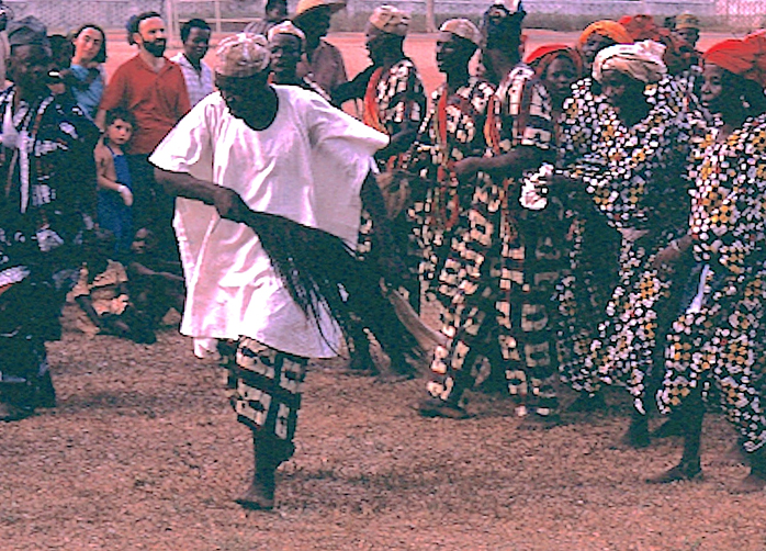 A photograph of a group of African dancers performing a traditional dance outdoors. The dancers are dressed in vibrantly patterned attire, with some wearing red head wraps. A man in the foreground, dressed in a white robe, is energetically swinging a horsetail whisk.