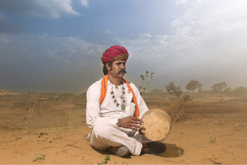 A photograph of a man dressed in traditional Rajasthani attire sitting on a sandy desert landscape under a cloudy sky. He wears a red turban, a white kurta, and a necklace with pendants. An orange scarf drapes over his shoulders. He holds a small drum in his right hand, with his left hand resting on his knee.