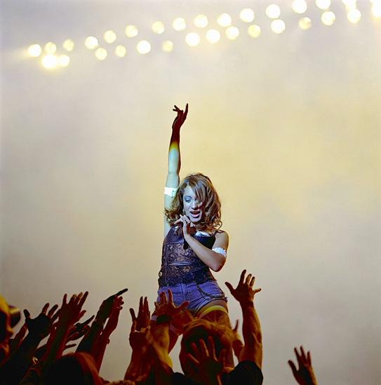 A dramatic stage performance photograph of a female pop singer in denim shorts and a lace top, raising one arm high while holding a microphone. Bright stage lights illuminate her from behind, and a crowd of fans reaches out towards her.