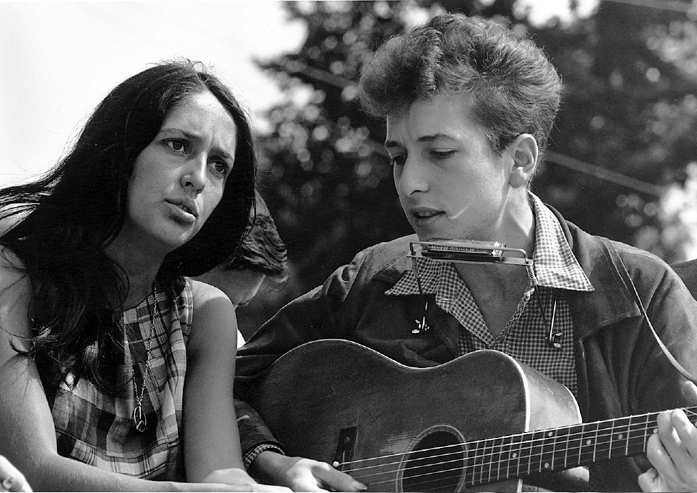 A black and white photograph of a man and woman playing music together outdoors. The man has curly hair, wears a checkered shirt under a jacket, and plays the guitar with a harmonica holder around his neck. The woman has long dark hair, wears a plaid dress, and sings with an intense expression.