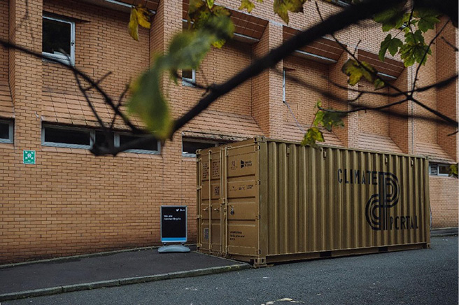 Khaki shipping container labelled “Climate Portal” beside an orange‑brick building; small pavement sign and overhanging branches in view.