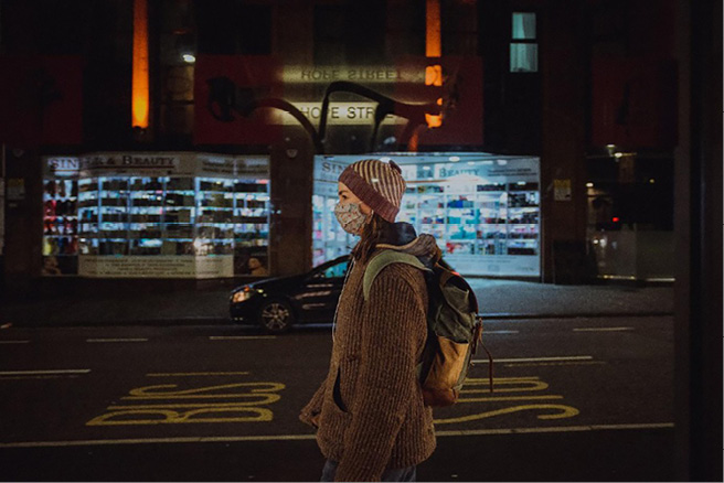 Woman in woollen hat, patterned face mask and rucksack walking along a night‑time city street past bright shop windows and bus‑lane markings.