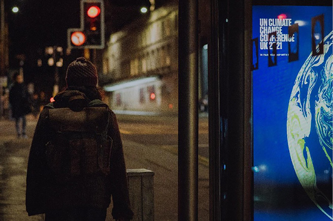 Back view of the same woman at a bus stop; a screen shows a ‘UN Climate Change Conference 2021’ poster while red traffic lights glow ahead in the dark street.