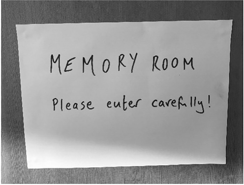 A handwritten sign reading ‘MEMORY ROOM’ and ‘Please enter carefully!’ is pinned to a wooden door, suggesting a reflective or sensitive space.