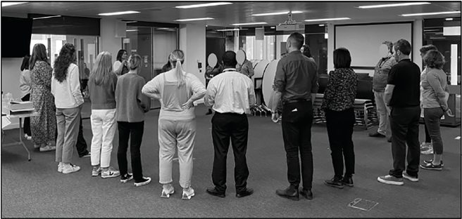 A group of adults standing in a semi-circle inside a modern room, appearing to participate in a workshop or training session.